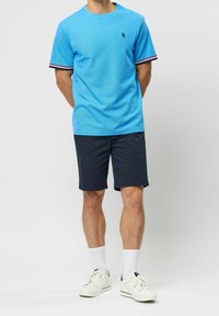 Jasnoniebieska koszulka z krótkim rękawem z kontrastowymi czarnymi, białymi i czerwonymi paskami na rękawach, zestawiona z granatowymi szortami i białymi sneakersami.