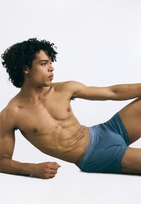 Zili kokvilnas bokseršorti ar mīkstu jostu, uz kuras ir "Calvin Klein" logo. Modelis ar īsiem cirtainiem matiem guļ profila pozā.
