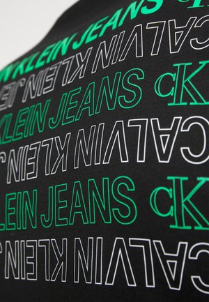 Tessuto nero con testo ripetuto in bianco e verde. Il testo recita "CALVIN KLEIN JEANS" in vari stili grafici e orientamenti.