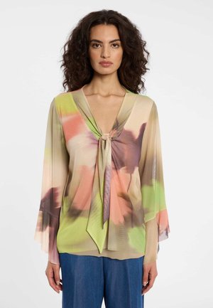 Vrouw met krullend haar draagt een doorschijnende blouse met bloemenprint, striksluiting en wijde mouwen, gecombineerd met een blauwe broek tegen een witte achtergrond.