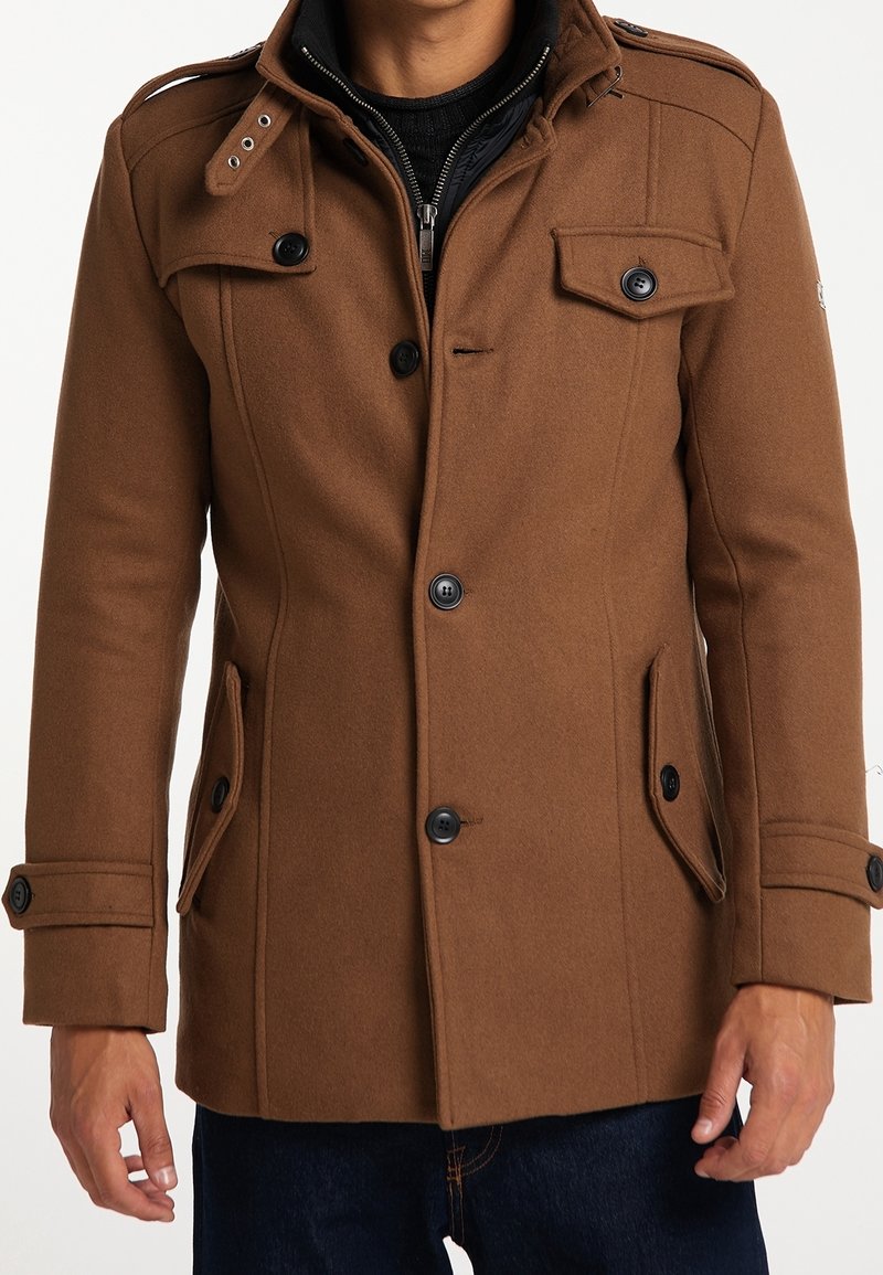 Hombre vestido con un abrigo de lana marrón con botones sobre un suéter negro con cremallera y jeans azul oscuro, de pie contra un fondo blanco.