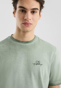 T-shirt à rayures vertes avec un col rond, présentant un logo noir sur la poitrine gauche. Fabriqué en tissu doux, texture lisse et coupe décontractée.