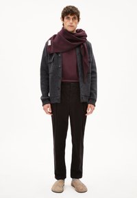 Donkergrijze wollen cardigan over een bordeauxrood shirt met lange mouwen, gecombineerd met zwarte broek en beige instapschoenen. Een bordeauxrode sjaal is om de nek gewikkeld.