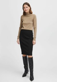Beige Rollkragenpullover aus weichem Material, kombiniert mit einem schwarzen, gerippten Bleistiftrock. Das Model trägt kniehohe schwarze Stiefel mit Absatz.