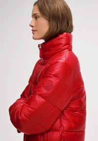 Czerwona kurtka puchowa z wysokim kołnierzem, błyszcząca powierzchnia, pikowana tekstura oraz naszywka z logo na lewym rękawku. Widoczny detal z suwakiem.