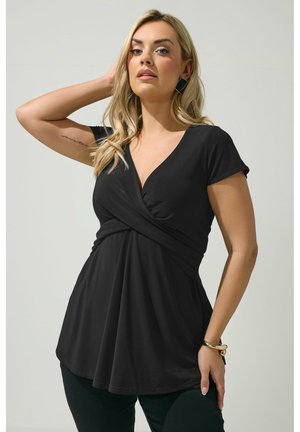 Blonde Frau mit lockigen Haaren trägt ein schwarzes Wickeloberteil mit V-Ausschnitt und kurzen Ärmeln, schwarze Hose, geometrische Ohrringe und ein goldenes Armband, posiert vor grauem Hintergrund.