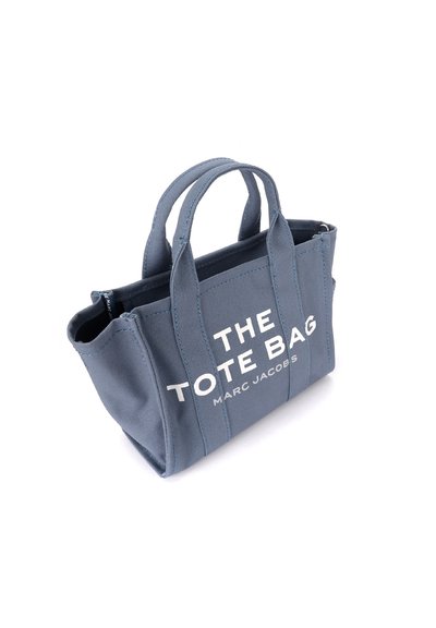 Sac fourre-tout en tissu bleu avec des poignées courtes, arborant un texte blanc qui indique "LE SAC FOURRE-TOUT MARC JACOBS." Forme rectangulaire, design robuste.
