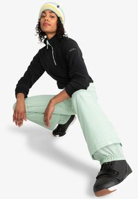 Sort fleece-pullover med halvlynlås, mintgrønne ski-bukser og sorte snowboardstøvler med brune såler. Beanie har grønne, gule og hvide striber.