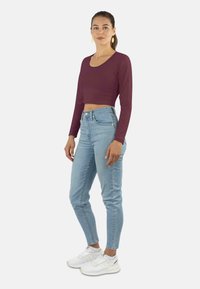 Burgunderrotes Langarm-Crop-Top, kombiniert mit hellblauen High-Waist-Jeans. Das Outfit wird durch weiße Sportschuhe ergänzt.