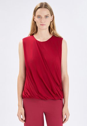 Donna con pelle chiara e lunghi capelli biondi, indossa una camicetta rossa senza maniche drappeggiata e pantaloni rossi coordinati, in piedi contro uno sfondo semplice.