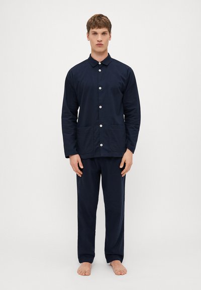 Navyblaue Baumwolljacke mit Kragen, Vorderknöpfen und zwei Taschen, kombiniert mit passenden, weit geschnittenen Hosen. Modell steht barfuß.