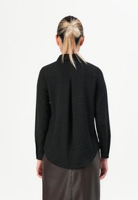 Blondide juustega naine, kes kannab musta pika varrukaga särki väikeste täppidega ja tumepruuni nahkpantsu, seisab valge taustaga seljaga camera poole.