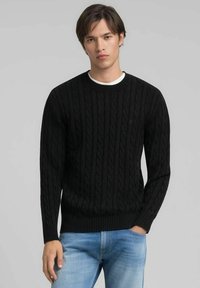 Pull noir en maille torsadée avec un col rond côtelé et des poignets, fabriqué dans un tissu épais et texturé. Porté avec un jean bleu clair.