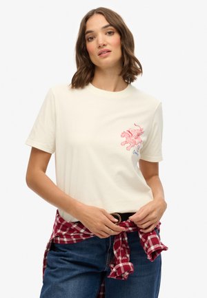 Crème T-shirt met korte mouwen, voorzien van een rode grafische afbeelding van een mythisch beest op de borst. Gecombineerd met blauwe spijkerbroek en een rode geruite blouse die om de taille is geknoopt.