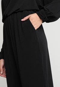 Svarta vida byxor med elastisk midja och sidofickor, i ett mjukt tyg med subtila veck för en avslappnad passform.