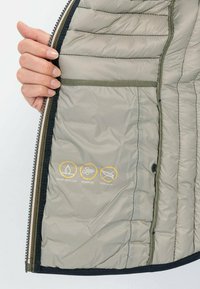 Lichte, waterafstotende grijze pufferjack met een gevoerde textuur en drie iconen die kenmerken aangeven: waterbestendig, winddicht, en vrij van dons.