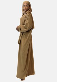 Lång, löst sittande tunika och matchande hijab i tannfärgad tyg, med en mjuk textur och flödande design. Skor i form av slip-on sandaler fulländar looken.