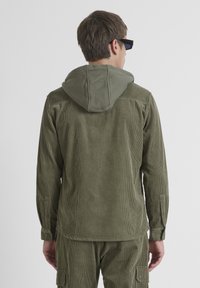 Chaqueta de pana verde oliva con capucha, que presenta franjas verticales, puños abotonados y una textura suave en la capucha.