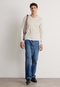 Maglione beige chiaro con scollo a v abbinato a jeans blu e sneakers nere. Il maglione presenta un orlo a coste ed è realizzato in un tessuto morbido.