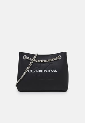 Černá kabelka Calvin Klein Jeans se stříbrným řetízkovým popruhem a vyraženým logem "CK" na přední straně.
