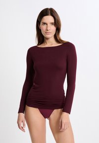 Donna con lunghi capelli castani che indossa un top aderente a maniche lunghe color bordeaux e biancheria intima abbinata, su uno sfondo chiaro e uniforme.