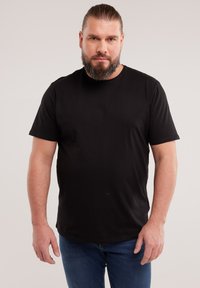 Habeme ja kinni seotud juustega mees, kes kannab lihtsat musta t-särki ja siniseid teksaseid, seisab neutraalse tausta ees.