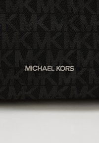 Czarna, teksturowana tkanina z wzorem logo. Srebrne logo "MICHAEL KORS" jest wyraźnie eksponowane na pierwszym planie.