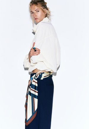 Vaaleahiuksinen nainen valkoisessa lyhyessä takissa, tummansinisiin housuihin pukeutuneena ja suuri raidallinen silkkinauhavyö, jolla siniset ja mustat sormukset kädessä rinnan päällä.