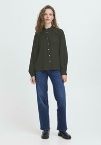 Femme portant une blouse noire à manches longues boutonnée, un jean droit bleu et des bottes noires à chevilles, se tenant contre un fond léger uni.