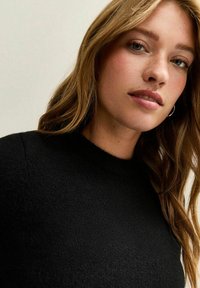 Schwarzer gestrickter Pullover mit hohem Kragen. Weiche Textur, figurbetontes Design. Die Haare sind wellig und umrahmen das Gesicht mit lockeren Locken. Dezente Creolen.