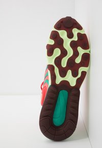 Sula av en färgglad sneaker med gröna, bruna och röda gummimönster, stående upprätt mot en vit hörnvägg och yta.