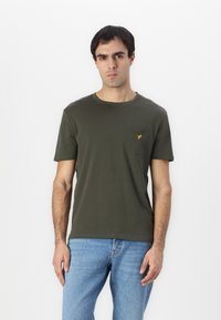 Camiseta de algodón de color verde oliva con cuello redondo, mangas cortas y un bolsillo en el pecho con un pequeño logo amarillo. Diseño simple, textura suave.