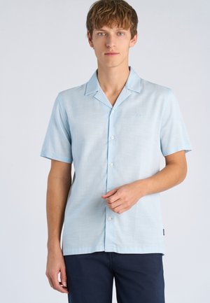 Junger Mann trägt ein hellblaues, kurzärmeliges Hemd mit Kragen und dunkle Hose, steht vor einem schlichten weißen Hintergrund.