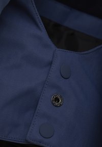 Mörkblå jacka i närbild som visar herringbone-textur, släta blå knappar och en dekorativ svart knapp med blommönster.