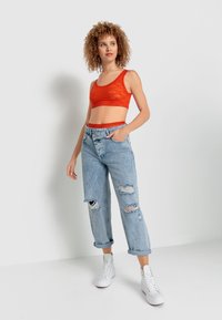 Rød blonde crop top med bølgede kanter, parret med lyseblå slidte jeans. Modellen har hvide sneakers på, hvilket fremhæver en afslappet æstetik.