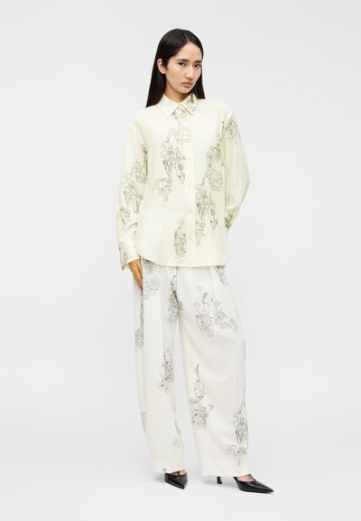 Πουκάμισο με μακριά μανίκια και κουμπιά, σε παλ κίτρινο χρώμα με λουλουδένια σχέδια γραμμής· συνδυασμένο με φαρδιά, λευκά παντελόνια με παρόμοια μοτίβα. Μαύρες γόβες.