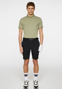 Jasnooliwkowa koszulka polo z zapięciem na guziki, zestawiona z czarnymi krótkimi spodniami, czarnym paskiem, białymi skarpetkami w paski oraz czarnymi butami sportowymi.