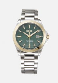 Montre en acier inoxydable avec un cadran à rayures vertes, des accents dorés, des aiguilles argentées, un affichage de la date et un bracelet à maillons. Étanche jusqu'à 50 m.