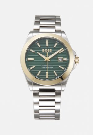 Stainless steel watch with a green striped dial, gold-tone accents, silver hands, date display, and a linked bracelet. Water resistant up to 50m.