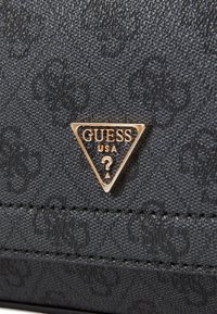Material texturizado negro con un logo triangular dorado "GUESS USA". Presenta detalles cosidos y un patrón sutil en el fondo.