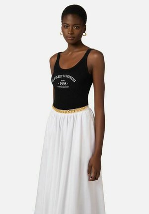 Donna con canotta nera con testo bianco, gonna bianca, cintura a catena dorata e orecchini a cerchio dorati su sfondo semplice.