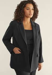 Blazer nero su misura con colletto a risvolto, maniche lunghe e tasche laterali. Tessuto liscio con una forma leggermente strutturata.