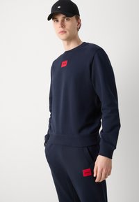Felpa blu marina realizzata in tessuto morbido, con una toppa rossa rettangolare sul petto. Abbinata a pantaloni jogger blu marina coordinati.