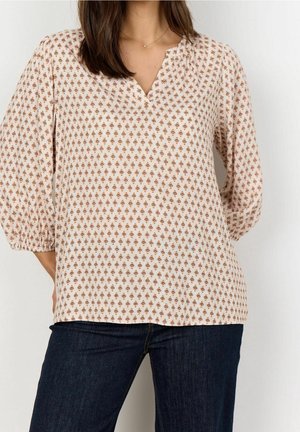 Vrouw draagt beige blouse met klein bruin geometrisch patroon en donkerblauwe spijkerbroek, staand met handen achter de rug.