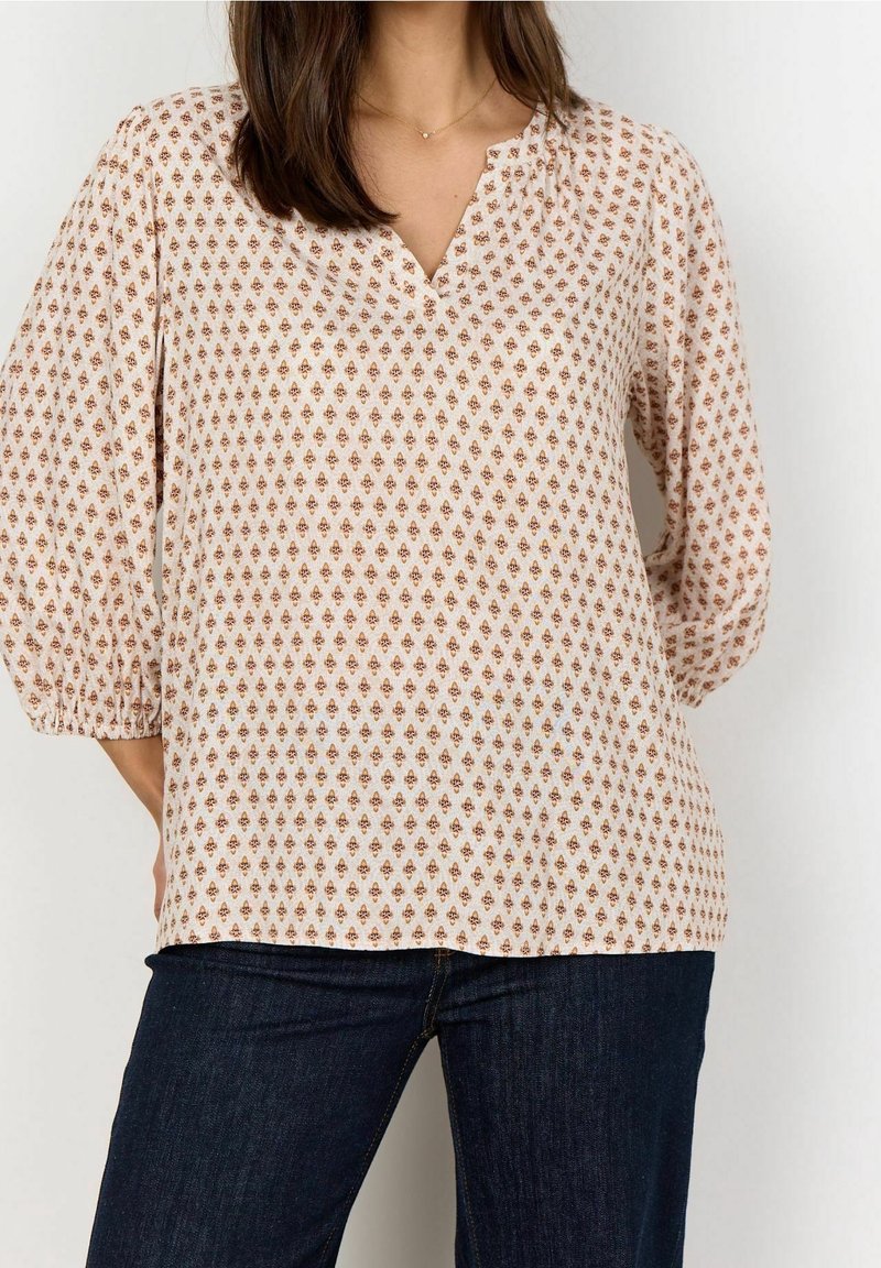 Femme portant un chemisier beige avec un petit motif géométrique marron et un jean bleu foncé, debout les mains dans le dos.