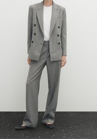 Costume gris croisé avec un tissu texturé, pantalon à jambes larges, et un t-shirt blanc ajusté en dessous. Associé à des chaussures noires basses.