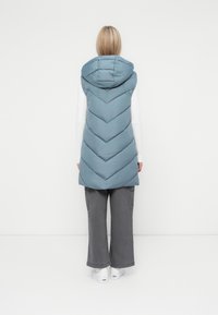 Svetlo modra podložena brezrokavnica s kapuco, z chevron quilted dizajnom, oblečena čez dolgi rokav in široke hlače.