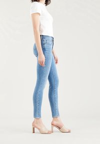 Jeans ajustados de tejido ligero en color azul claro con una cintura alta, que presentan un diseño de cinco bolsillos y un ligero desgastado, combinados con mulas de tacón abierto en color beige.