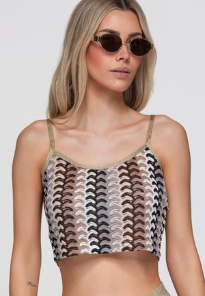 Ung kvinde med blondt hår iført mørke solbriller og en ærmeløs crop top med bølgede striber i neutrale toner.