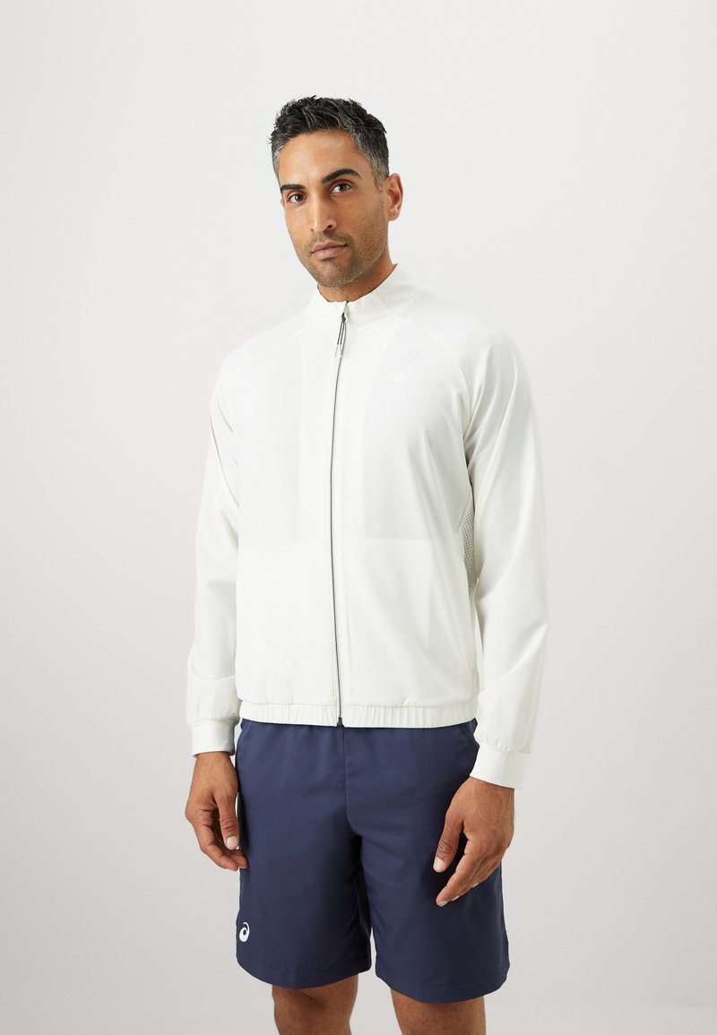 Helles Sportjackett mit hohem Kragen, Frontreißverschluss und seitlichen Mesh-Einsätzen, kombiniert mit navyblauen Shorts mit Logo.